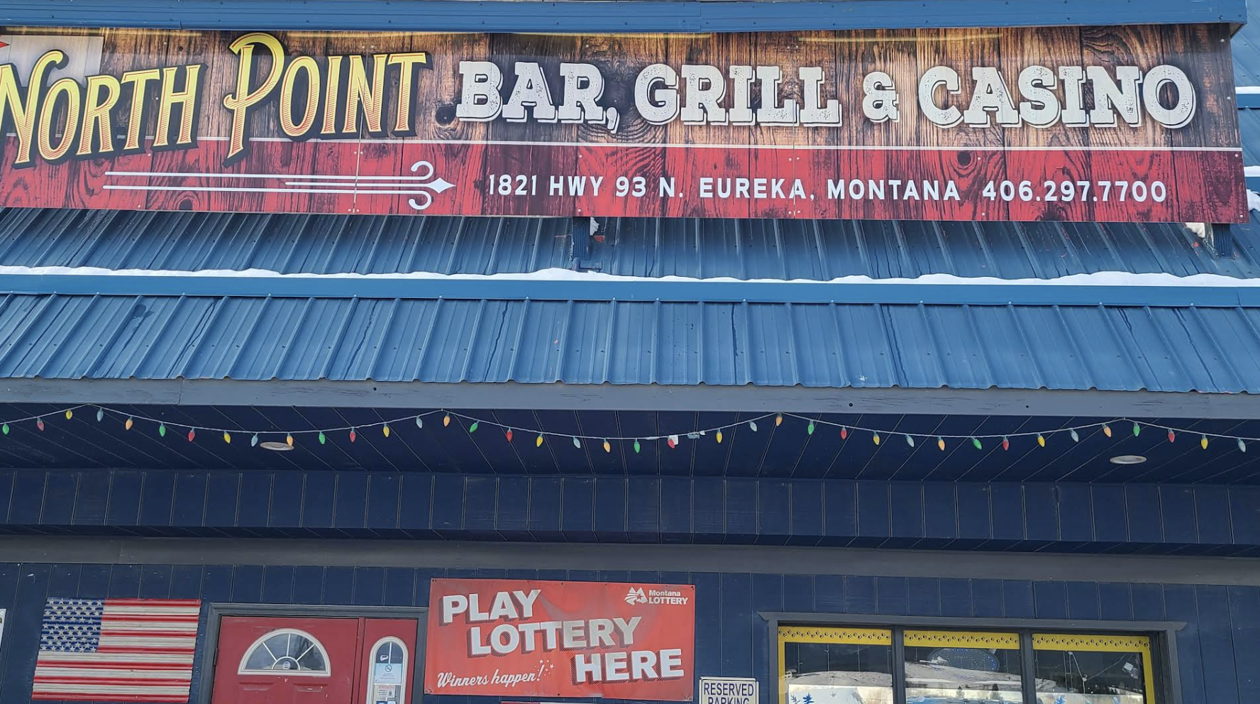 Sign for North Point Bar, Grill & Casino, located at 1821 Highway 93 North in Eureka, Montana, with a phone number 406-297-7700, the building has a blue roof and string lights overhead, and a red door with a window. There is also a Montana Lottery sign and a portion of an American flag display.
