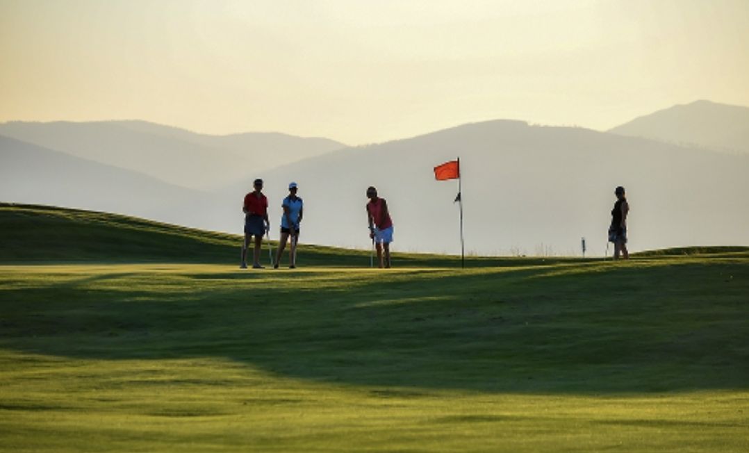 Four people standing on a golf course green near a flag, with mountains in the background during sunset or sunrise.
