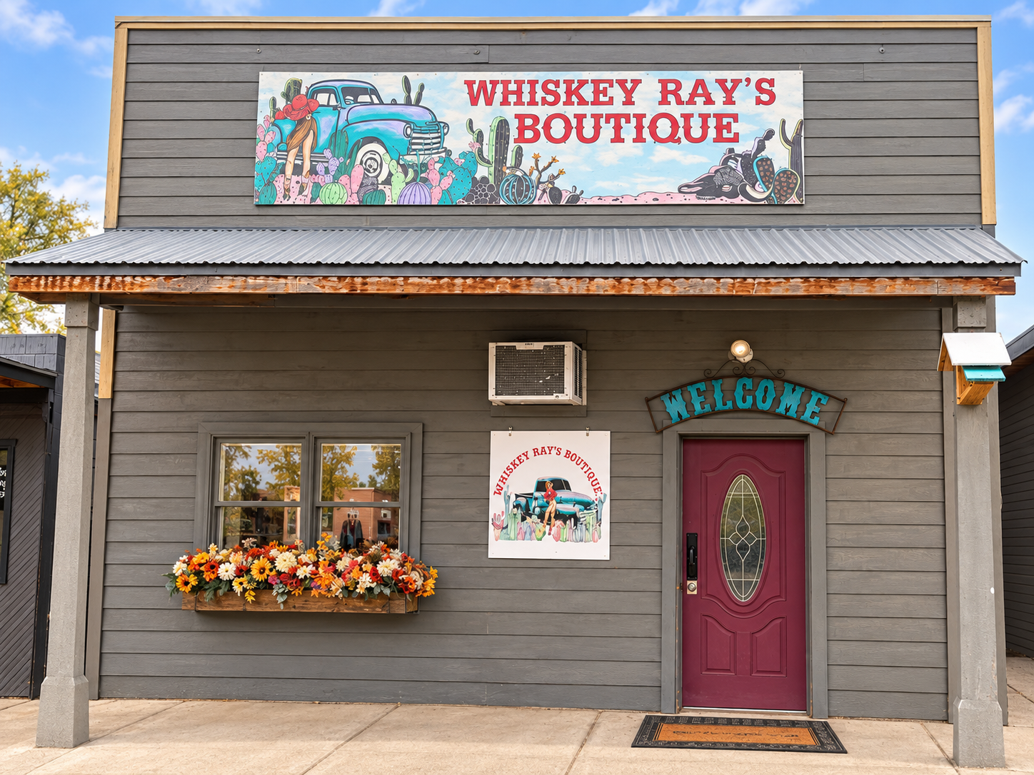 The exterior of a boutique store named 'Whiskey Ray's Boutique' with a large colorful sign on top showing a vintage truck, cacti, and desert scenery, against a partly cloudy sky. The storefront has a window box filled with orange, yellow, red, and white flowers, a maroon door, a welcome sign, and an air conditioning unit above a smaller sign with the same vintage truck illustration.