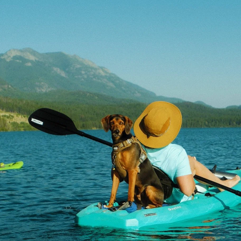 Person in a light blue shirt and straw hat kayaking on a lake with a dog sitting on the kayak, mountains in the background.