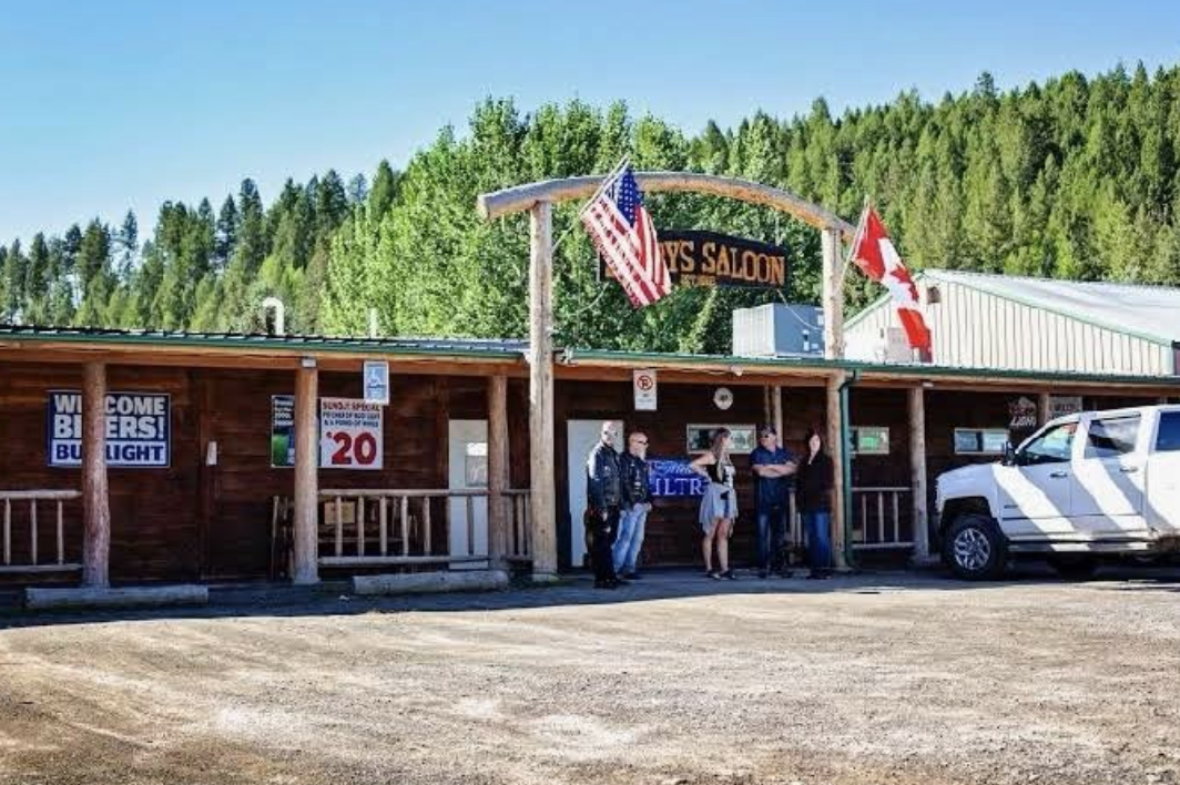 A rustic wooden building with a sign reading 'Lynch's Saloon' and American and Canadian flags, with a group of five people standing outside near a parked white truck, set against a background of green trees and a clear blue sky.