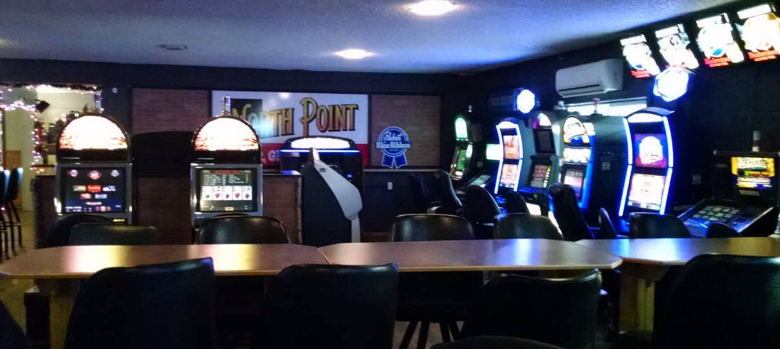 Interior of a casino or gaming arcade with multiple slot machines with illuminated screens, a row of video poker machines, and a table with chairs in the foreground.