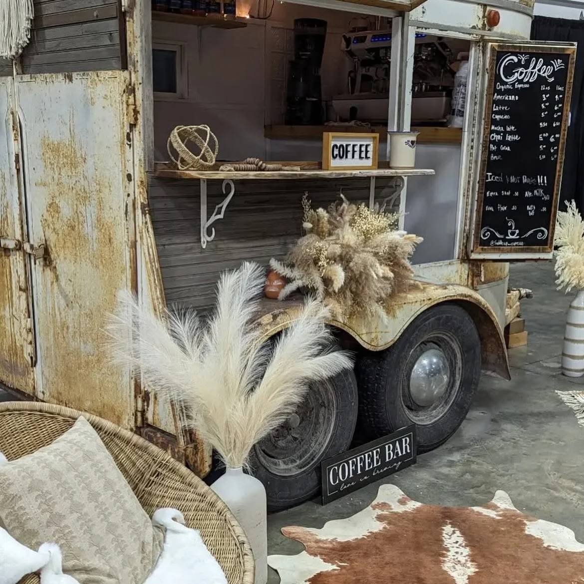 A rustic mobile coffee stand on a trailer with a weathered yellow exterior, decorated with dry pampas grass and neutral-toned accents. A blackboard menu lists various coffee drinks, and a small sign reads 'COFFEE'. There are rattan furniture and decorative pillows nearby, and a cowhide rug on the floor.