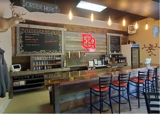 Bar area inside a restaurant or brewery with a wooden counter, black chairs, a deer mounted on the wall, and a neon sign with the letters 'BC'.