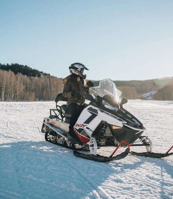 A person wearing a helmet and winter clothing sitting on a snowmobile in a snowy landscape with a forested hillside in the background.