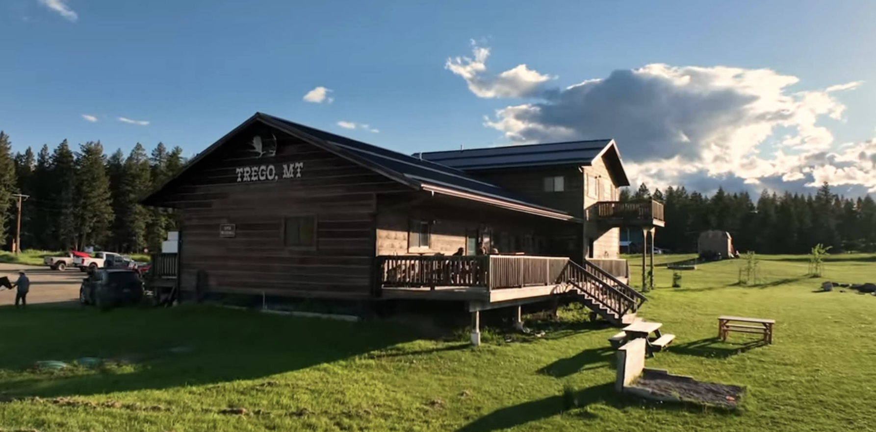 A wooden building with the words 'Trego, MT' on it, surrounded by green grass, trees, and a clear blue sky with clouds.
