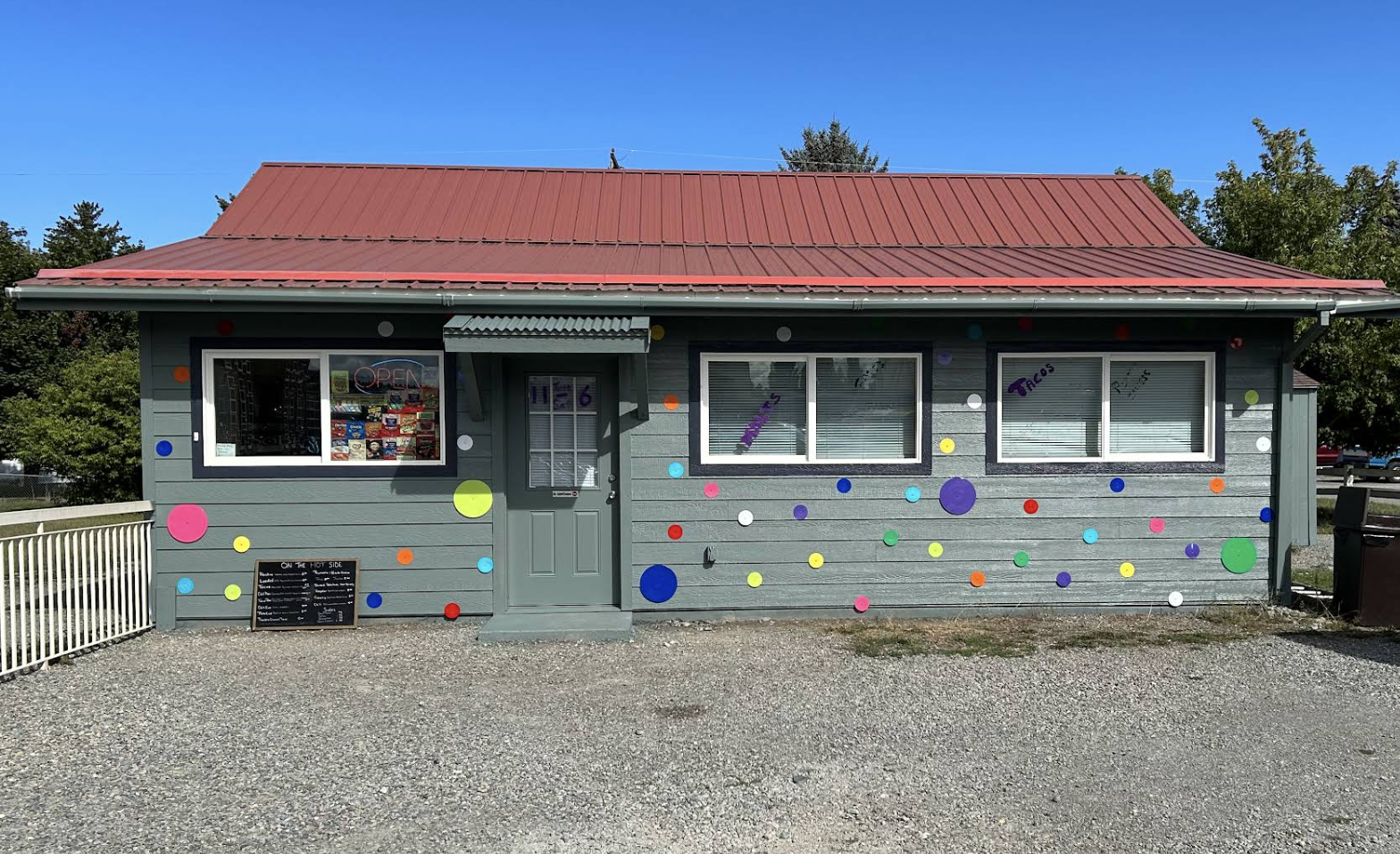 Small shop building painted gray with multicolored polka dots, a red metal roof, and three windows with blinds, one showing drinks on the glass. There is a boarded entrance door with a small awning above it, and a chalkboard menu outside. A neon sign in the window reads 'OPEN'.