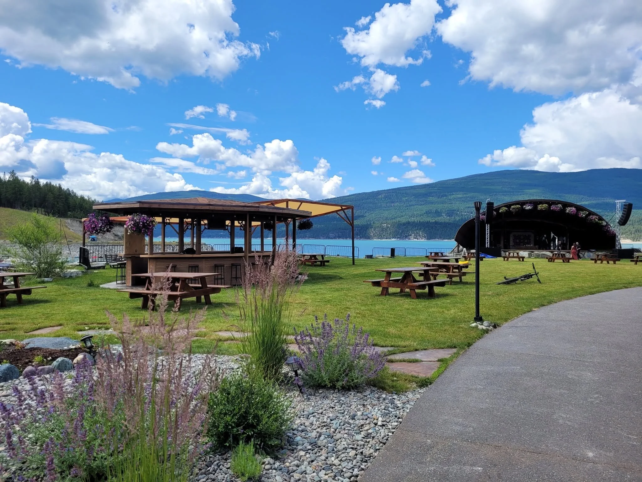 A scenic outdoor setting near a lake with green grass, flowering plants, picnic tables, a small pavilion, and a stage, with mountains and a partly cloudy sky in the background.
