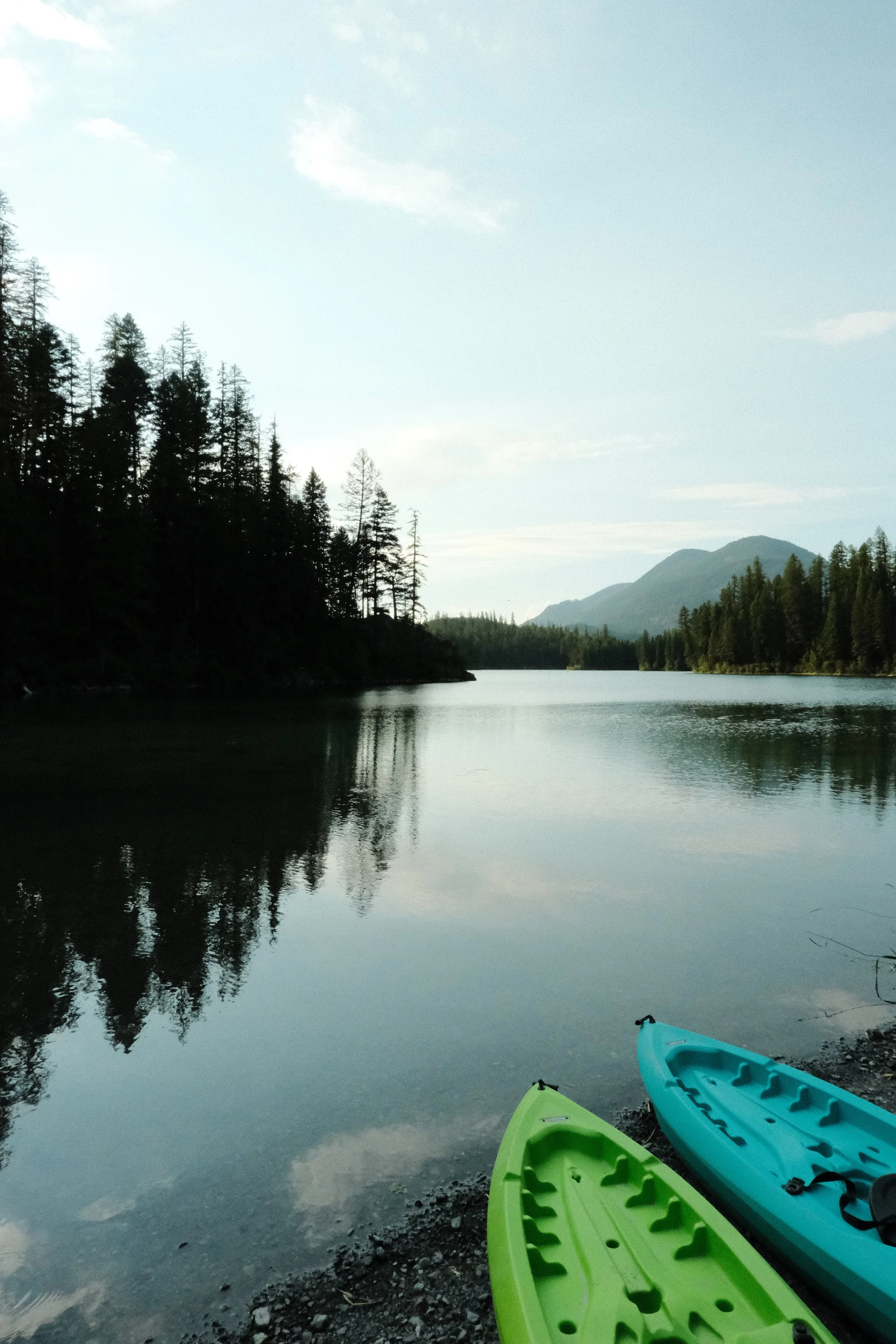 Colorful kayaks on the rocky shore of a calm lake, surrounded by trees and mountains, under a partly cloudy sky.