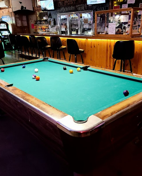 Pool table in a bar with balls on the table, a bar counter with stools, and a mirror behind the bar showing beer taps and bottles.