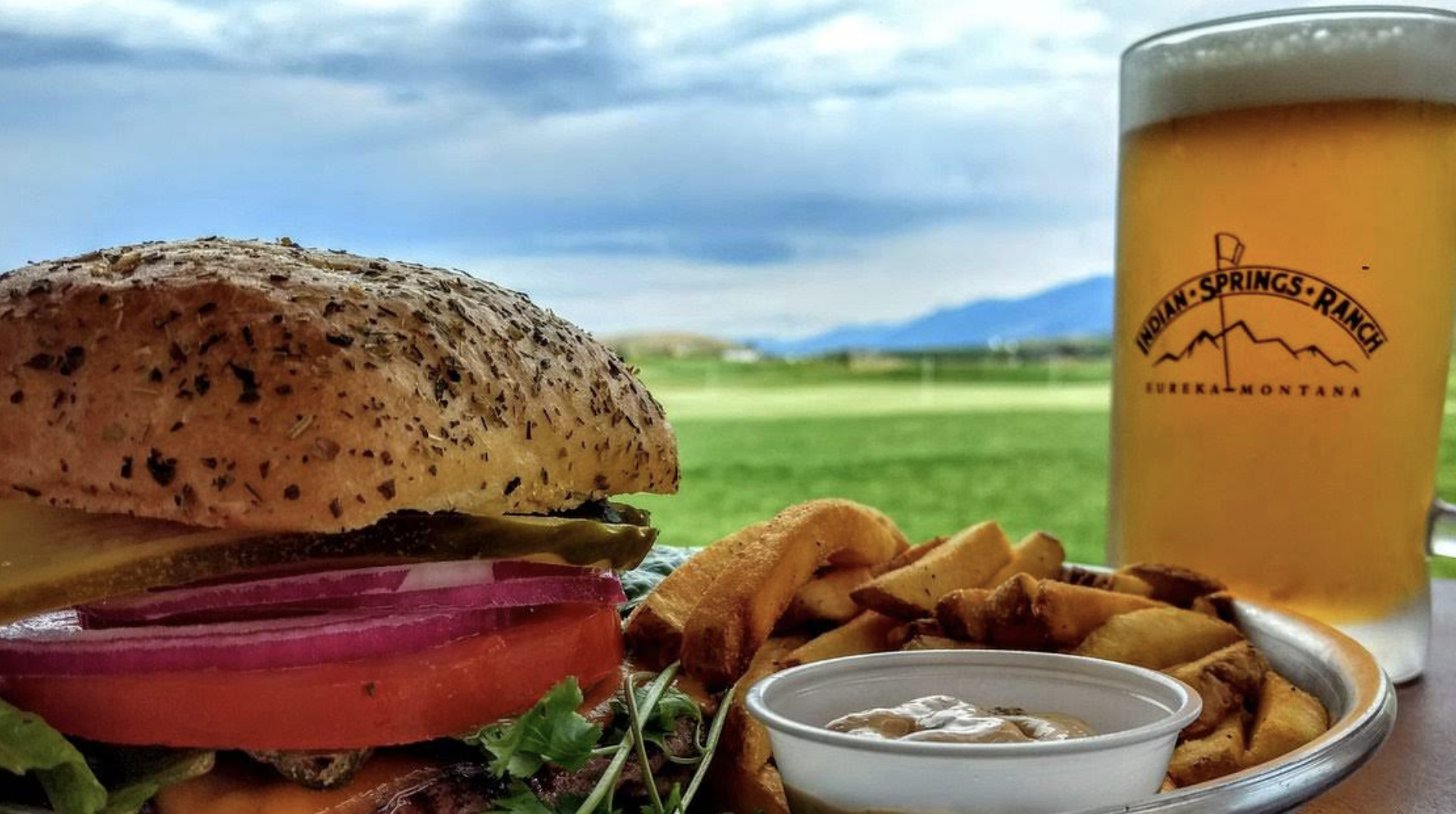Close-up of a burger with vegetables, fries, a small cup of dipping sauce, a glass of beer, and a scenic outdoor background with a field and mountains.