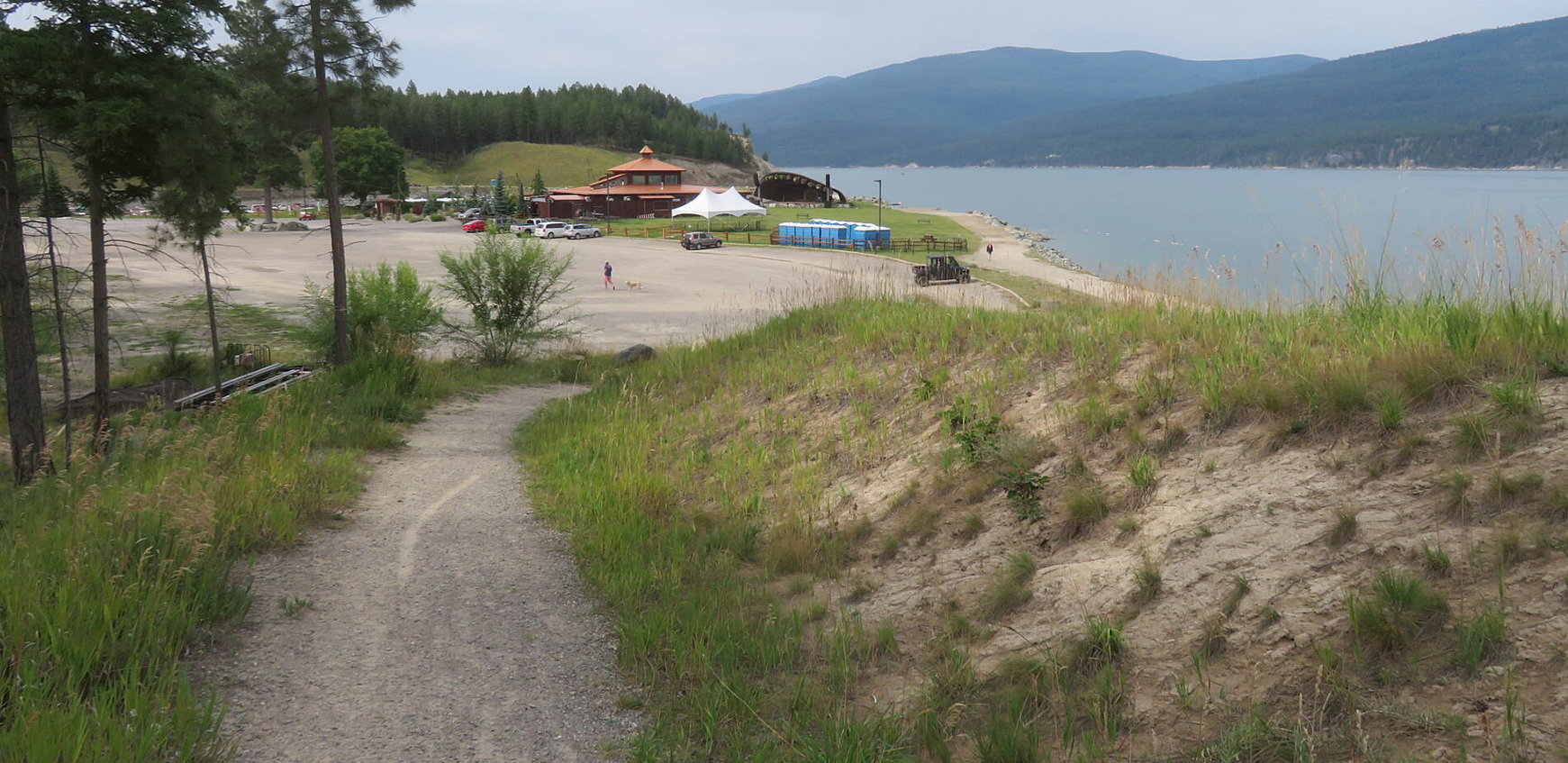 A gravel path leading down a sandy dune towards a lakeside with a building, tents, and parked cars, surrounded by trees and hills in the distance.