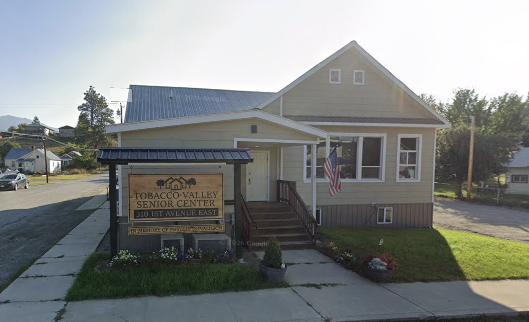 The image shows a building with a sign in front that reads "Tobacco Valley Senior Center, 310 1st Avenue East, In Memory of Phyllis Donaldson." The building is a two-story house painted in light yellow with white trim and a dark roof. An American flag is hanging near the front windows. The area surrounding the building includes a sidewalk, small flower beds, and a grassy lawn. In the background, there are other houses, trees, and a hilly landscape.