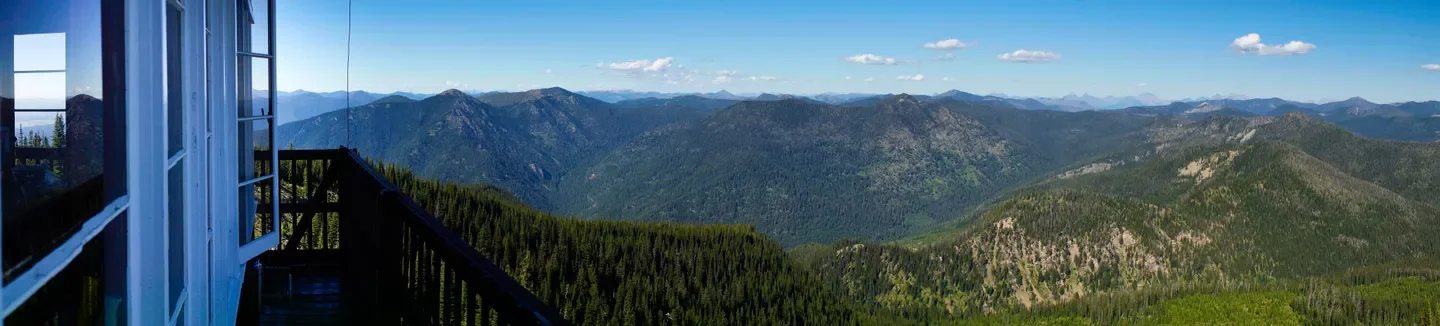 View from a mountain cabin deck showing a mountain range with lush green forests and clear blue sky.