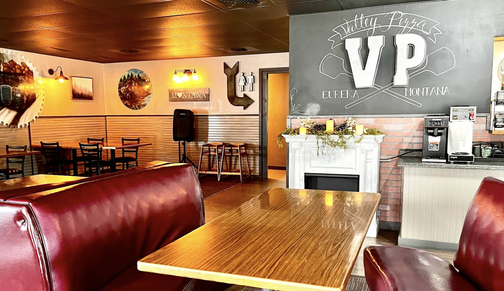 Cozy restaurant interior with wooden tables and red leather booths, wall art featuring Montana scenery, a chalkboard sign with 'Statley Pizza VIP' and 'Eureka Montana,' a fireplace decorated with greenery and candles, and a coffee station.