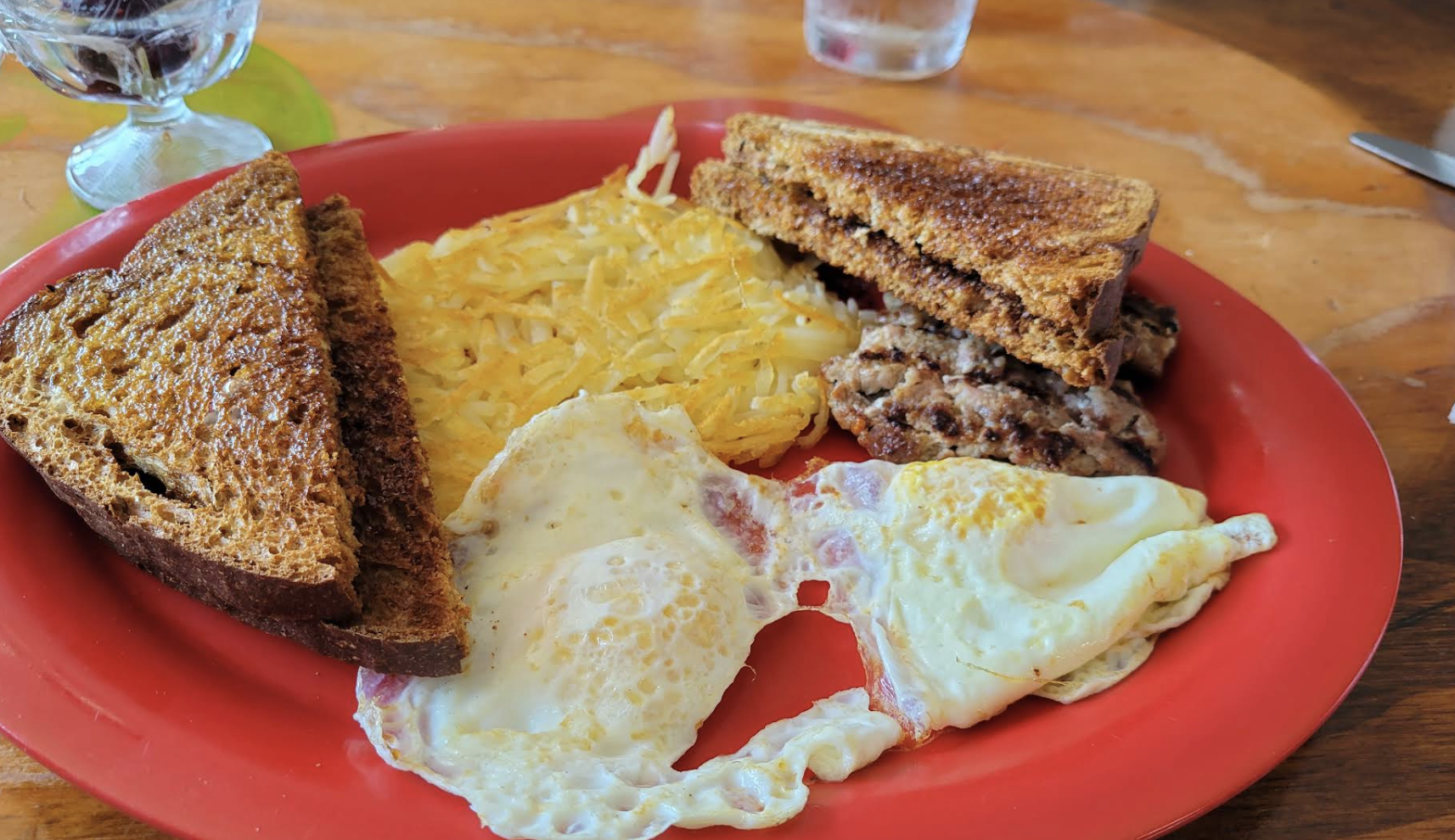 A breakfast plate with fried eggs, hash browns, toast, and chocolate chip pancakes on a red plate.