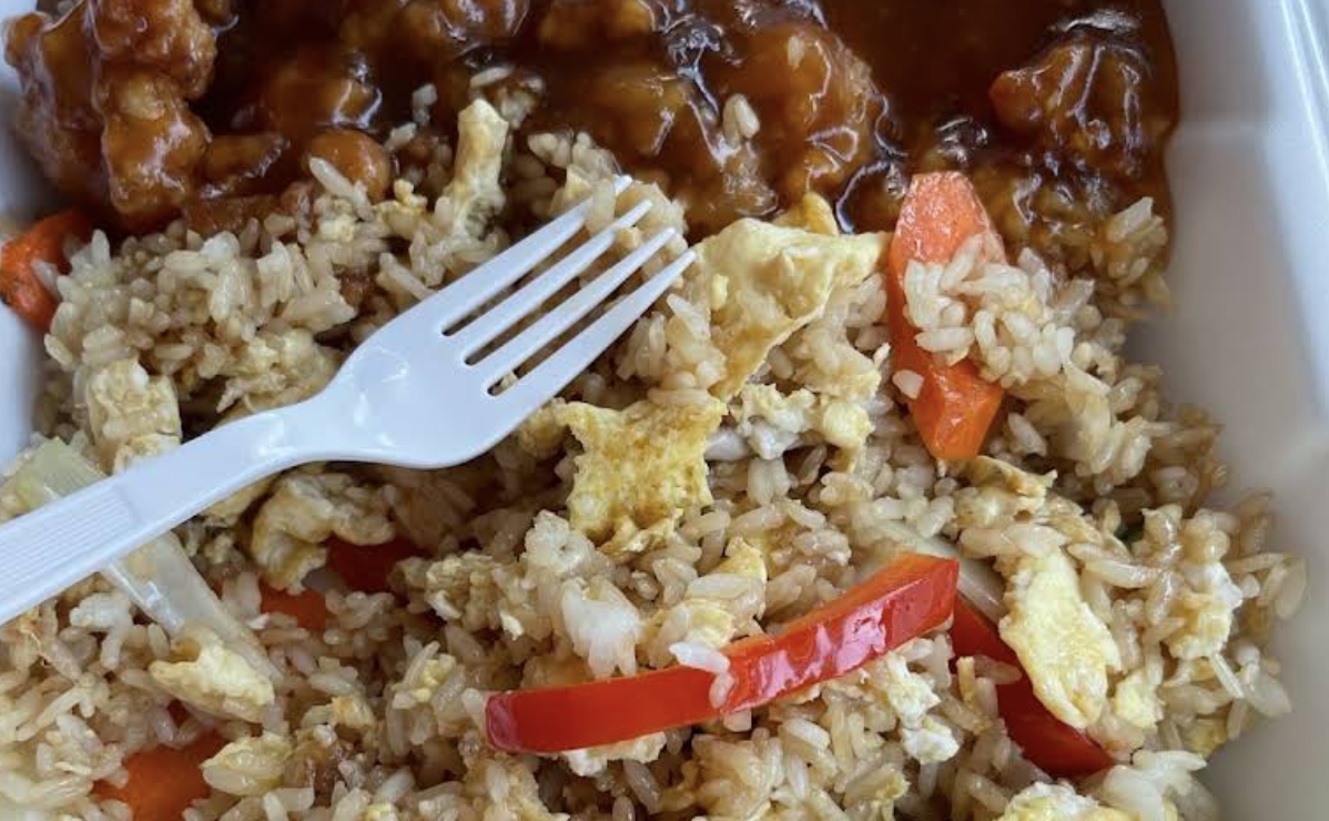 Close-up of a takeout container with fried rice, scrambled eggs, red bell peppers, and a serving of sweet and sour chicken in sauce, with a white plastic fork resting on the rice.