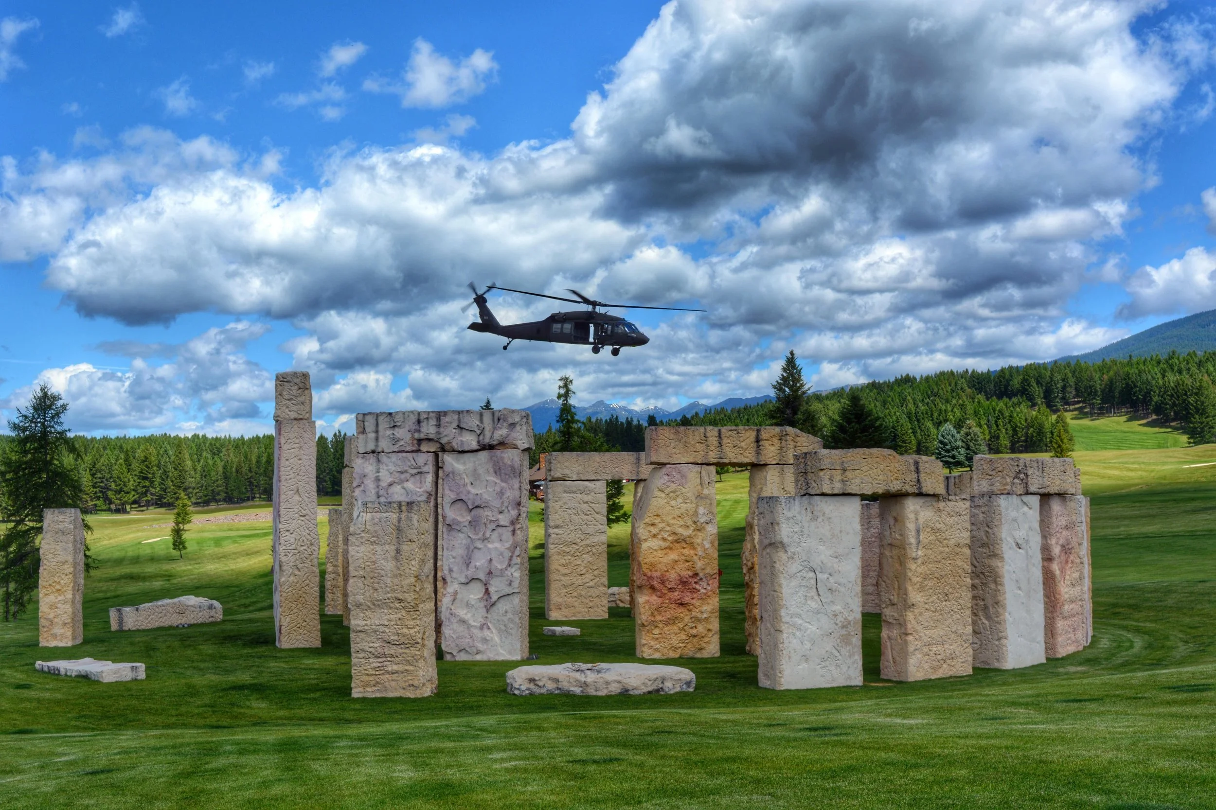 A helicopter flying over a stone structure on a grassy field with a forested landscape and mountains in the background under a partly cloudy sky.