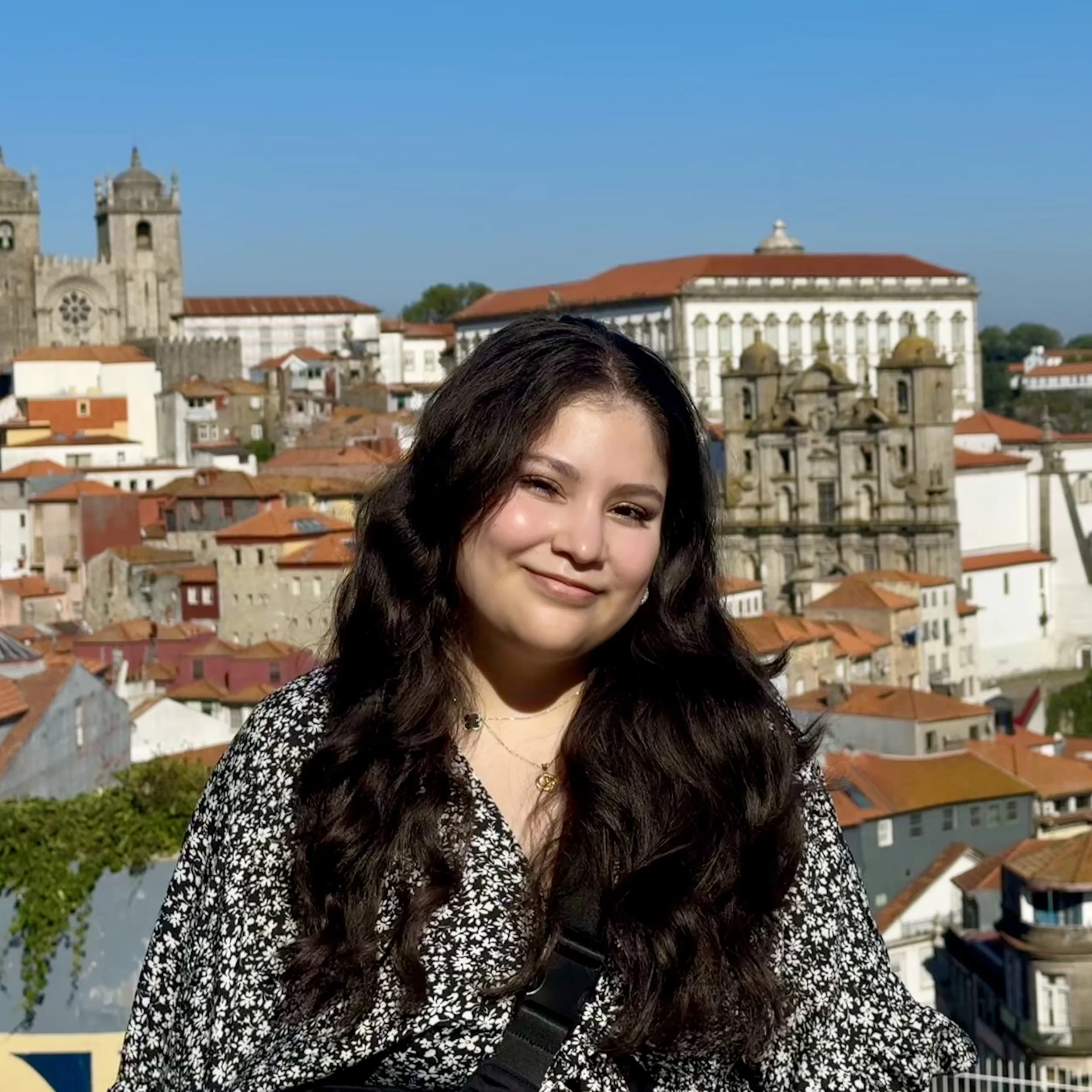 Partial view of a smiling woman with long dark hair in front of historic buildings with red-tiled roofs and a church with two towers under a clear blue sky.