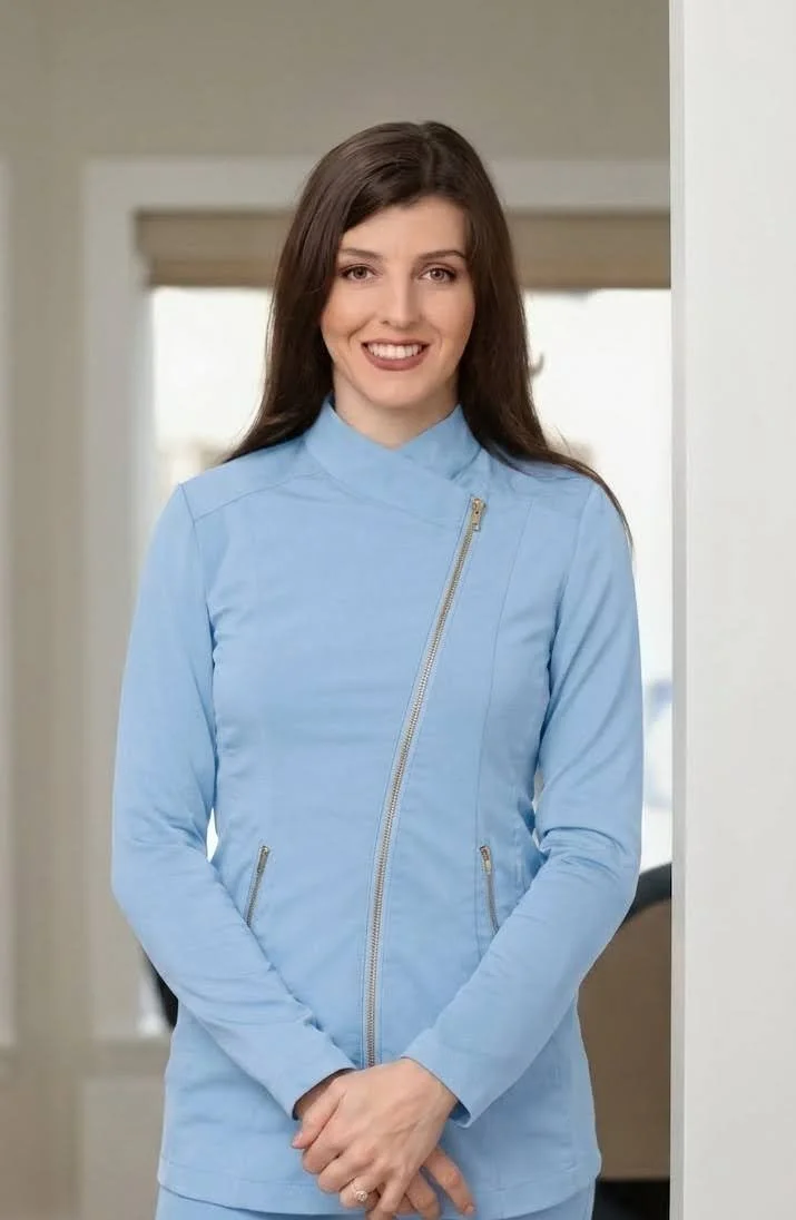 A woman with long brown hair smiling, wearing a light blue jacket with zippers, standing indoors near a wall.