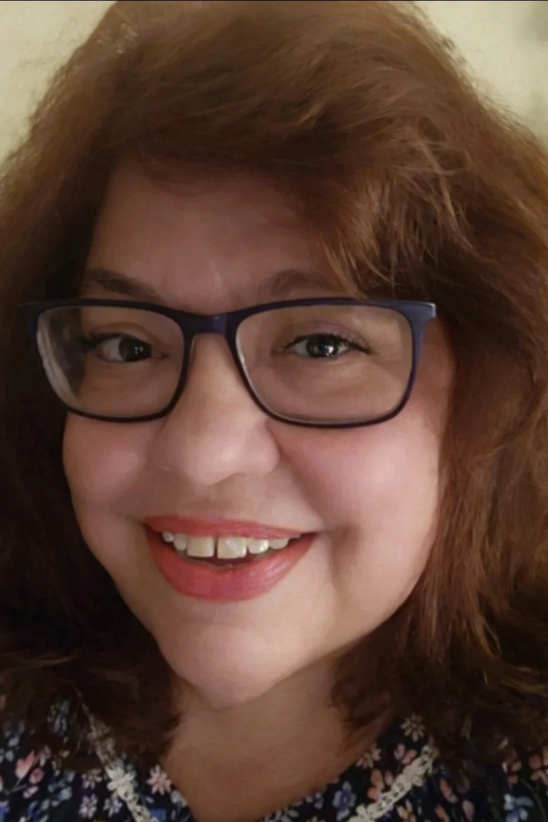 Close-up of a woman with reddish-brown hair wearing glasses and a floral top, smiling.