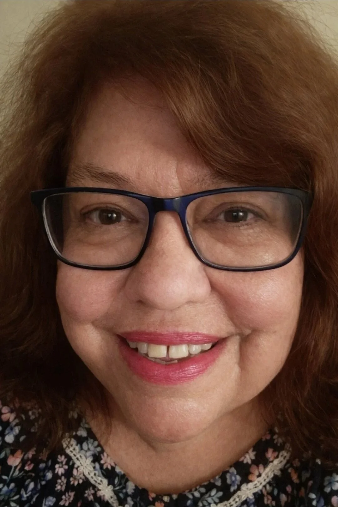 A close-up of a smiling woman with red hair, glasses, wearing a floral top.
