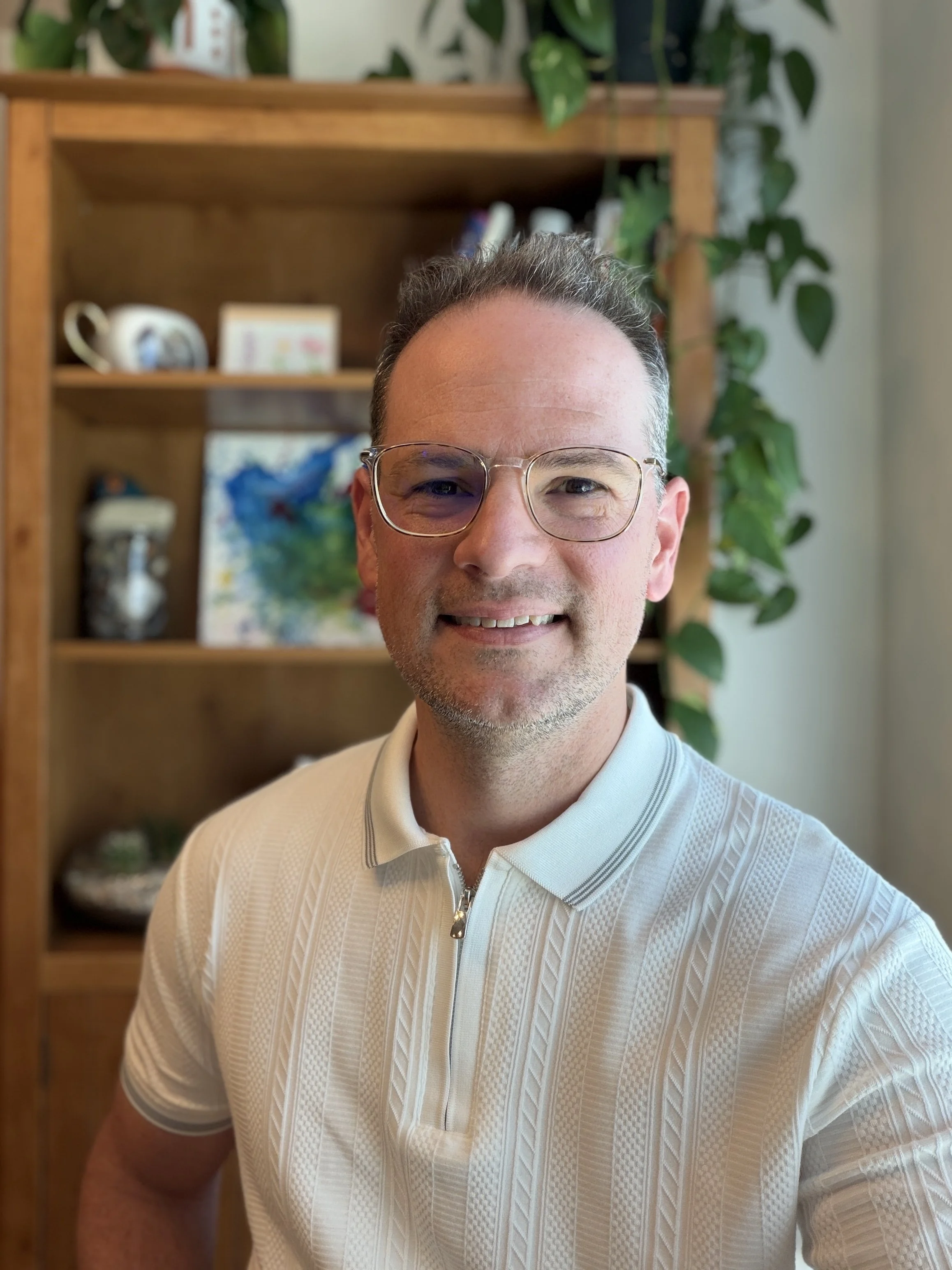 Portrait of a smiling man wearing glasses and a white textured polo shirt, standing in front of a wooden shelf with potted plants, books, a mug, and decorative items.