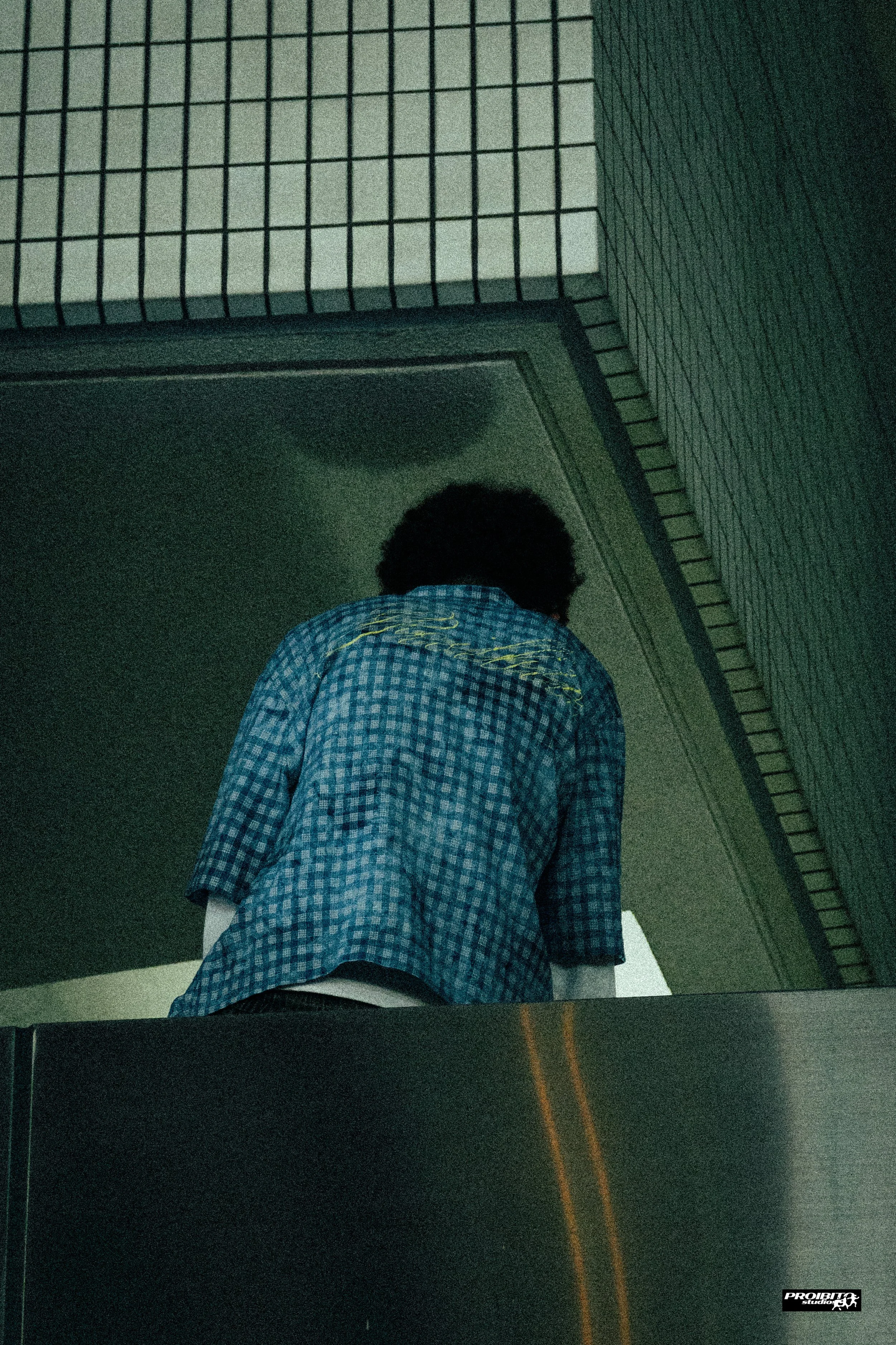 A person with curly black hair wearing a blue checkered shirt partially visible from the back, standing at a window or mirror in a dimly lit space.