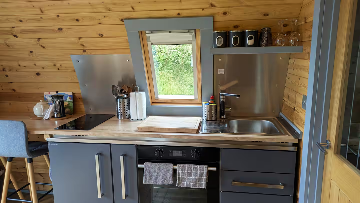 Small modern kitchen with wood-paneled walls, gray cabinets, and a countertop with a window in the center showing greenery outside. There is a stovetop, a sink, and various kitchen items including a cutting board, paper towel holder, and spice contai