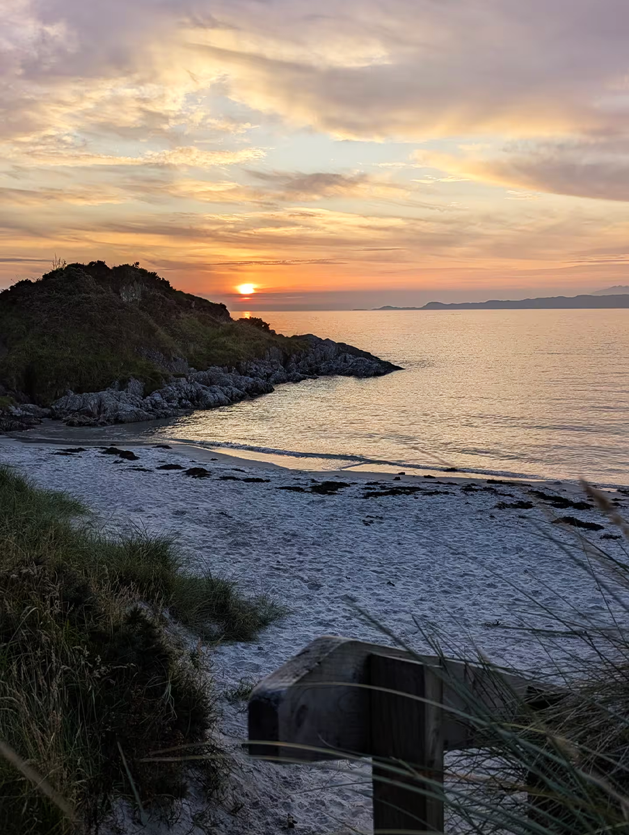 A beach at sunset with a rocky headland and calm ocean waters, with a partly cloudy sky and distant landmasses.