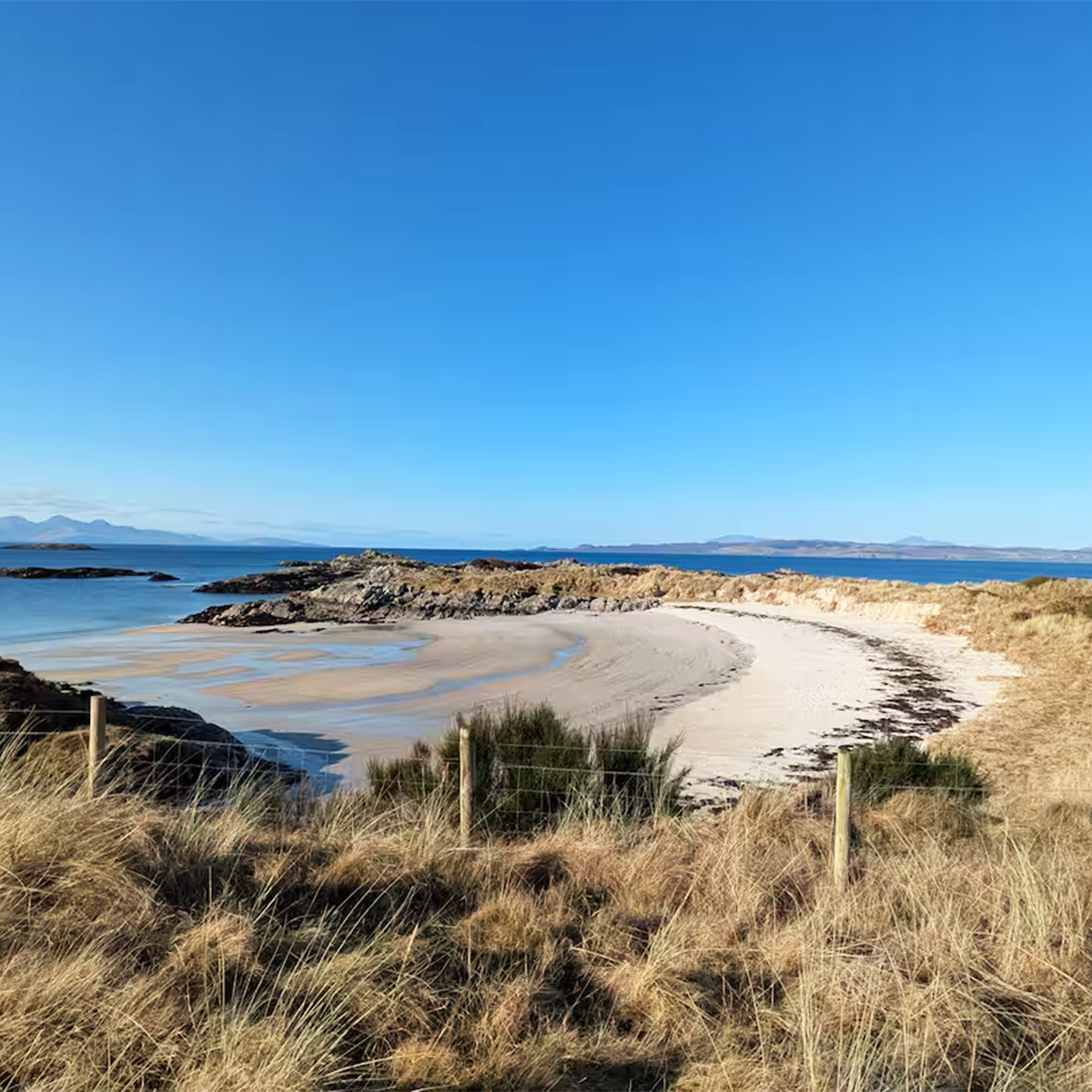 A sandy beach with grassy dunes in the foreground, rocky formations along the shoreline, calm blue water, and a clear blue sky with distant mountains in the background.