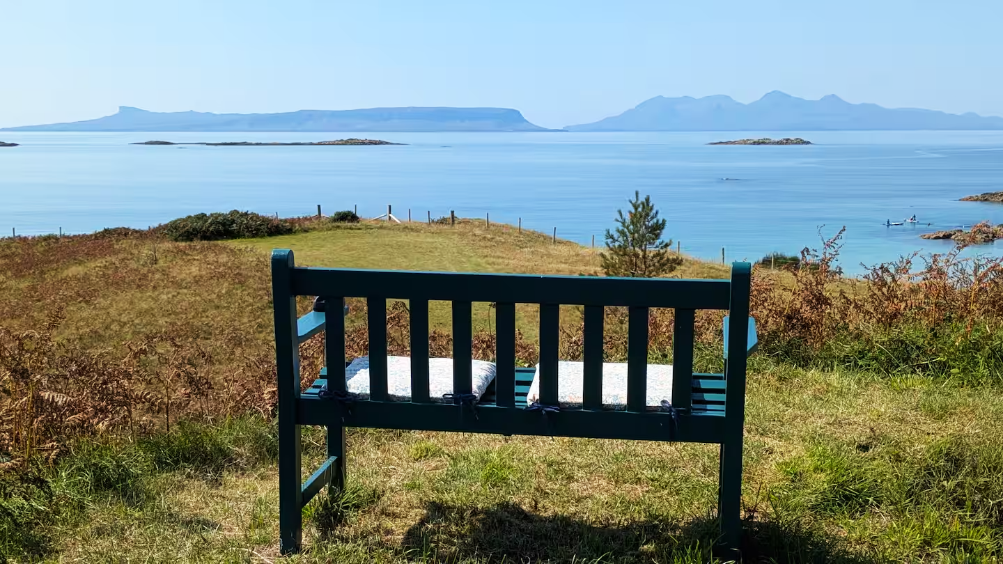 A dark green bench on a grassy hill overlooking a large body of water with small islands and mountains in the background.