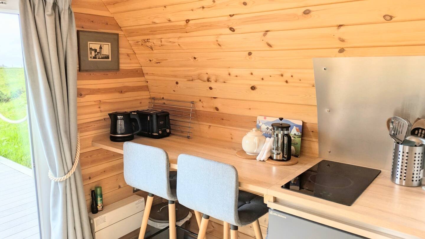 Kitchen area with a wooden wall, a counter with a coffee maker, toaster, kettle, and utensils, and two light gray chairs facing the counter.
