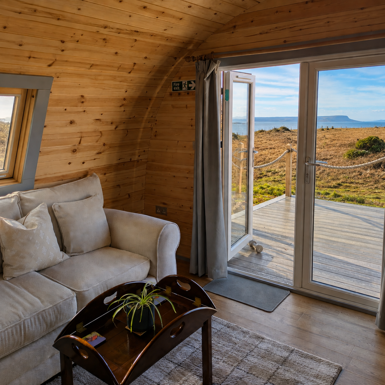 Interior view of a cozy wooden cabin living room with a cream couch, small wooden coffee table with a potted plant, and a sliding glass door revealing a deck and scenic landscape with grass, water, and hills in the background.