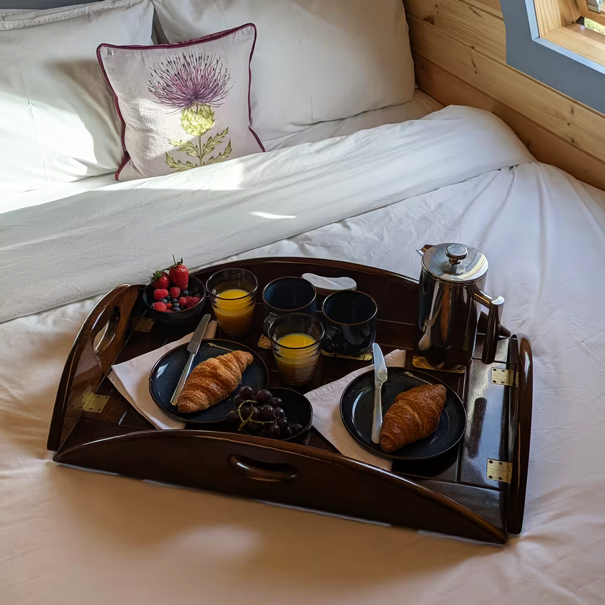 Breakfast tray on a bed with croissants, fresh berries, orange juice, coffee, and a teapot, in a cozy bedroom with a floral pillow and wooden wall.