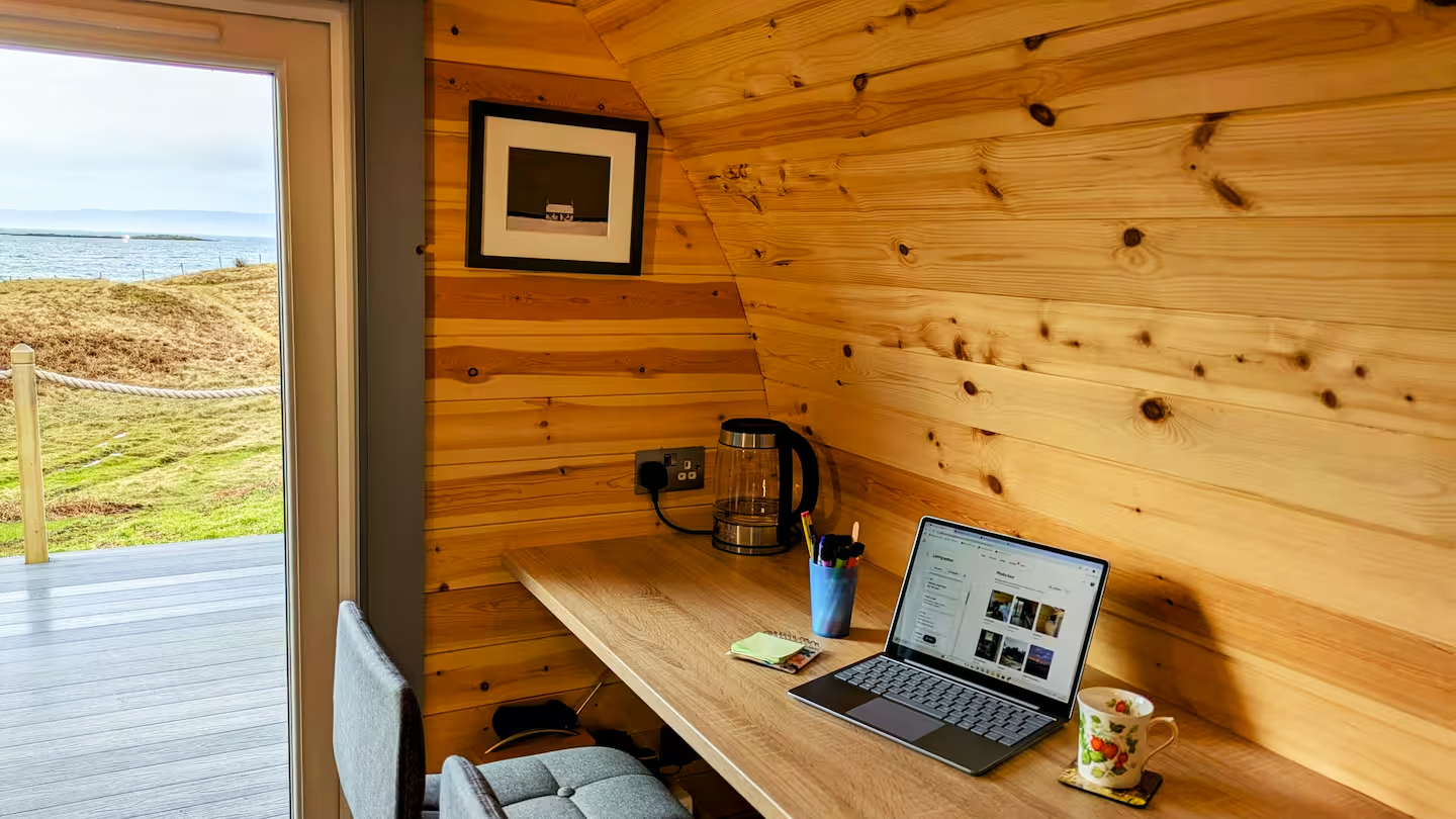 Wood-paneled home office with a desk outside near a window showing a lakeside view, contains a laptop, a coffee mug, a container with pens, a notebook, a kettle, and a framed picture on the wall.