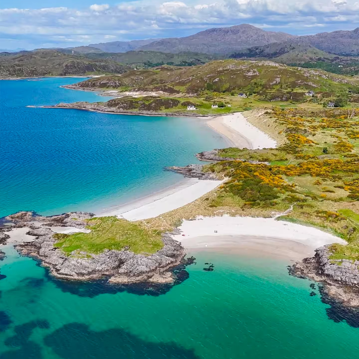 Aerial view of a pristine beach with white sand, surrounded by rocky cliffs and lush green hills, with blue ocean water.