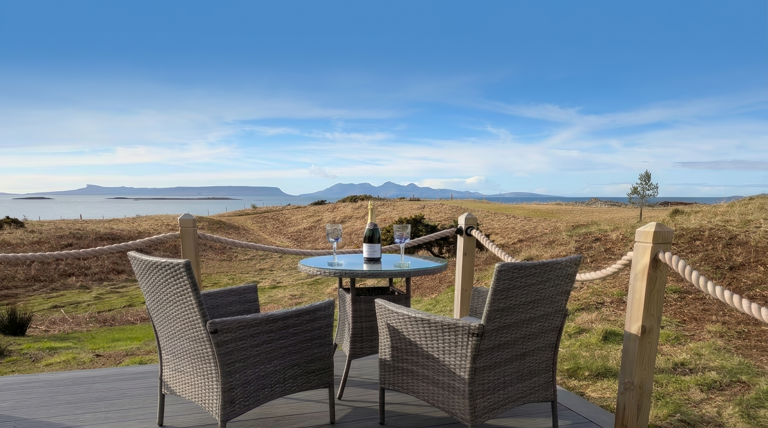 A table set for two with a bottle of champagne and two glasses overlooking coastal hills and mountains under a blue sky.