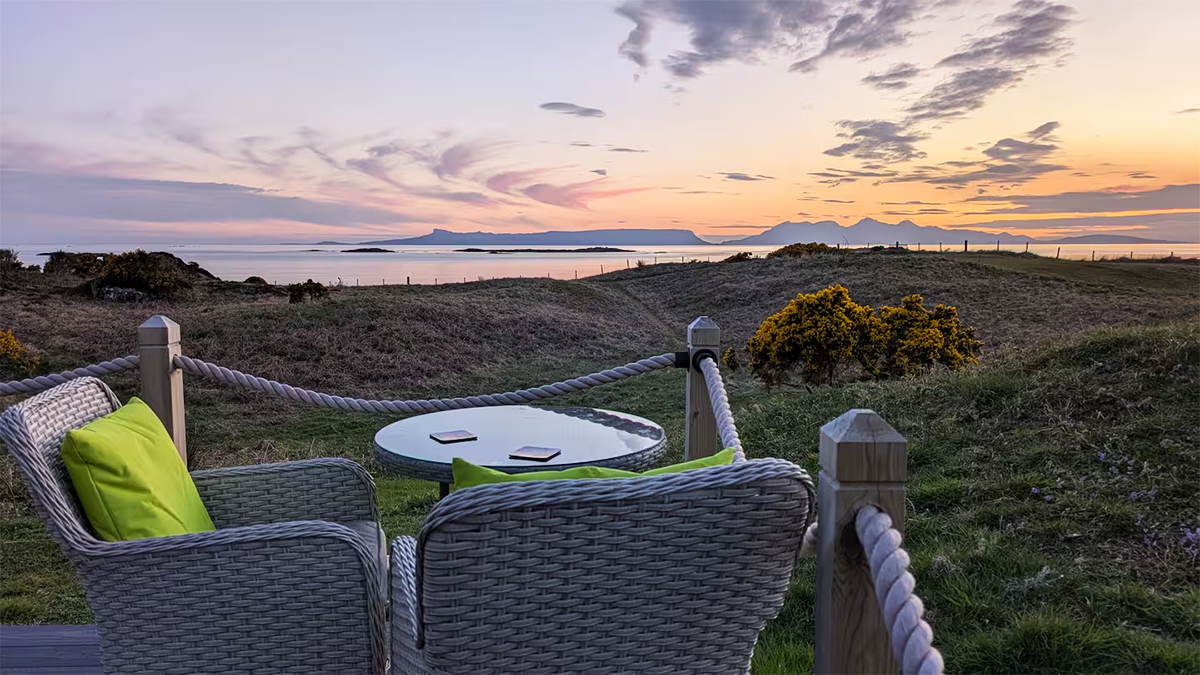 Outdoor seating area with two wicker chairs with lime green pillows and a glass table, overlooking a grassy landscape with hills, water, and islands during sunset.