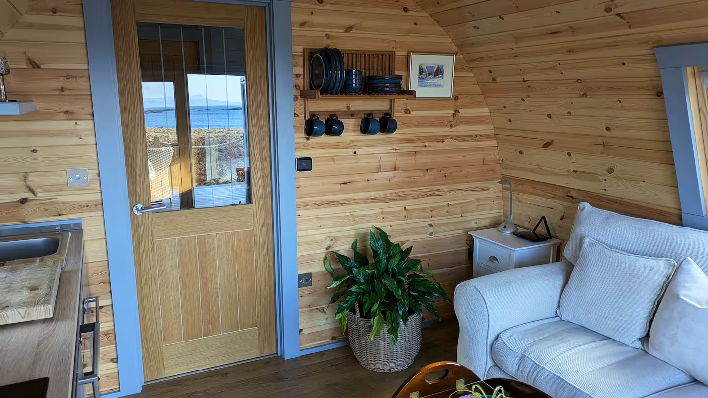 A cozy living space with wooden walls and ceiling, a white sofa with cushions, a small white side table with a lamp, a green potted plant in a wicker basket, a wooden door with glass panels leading outside, and a shelf with plates, cups, and a framed