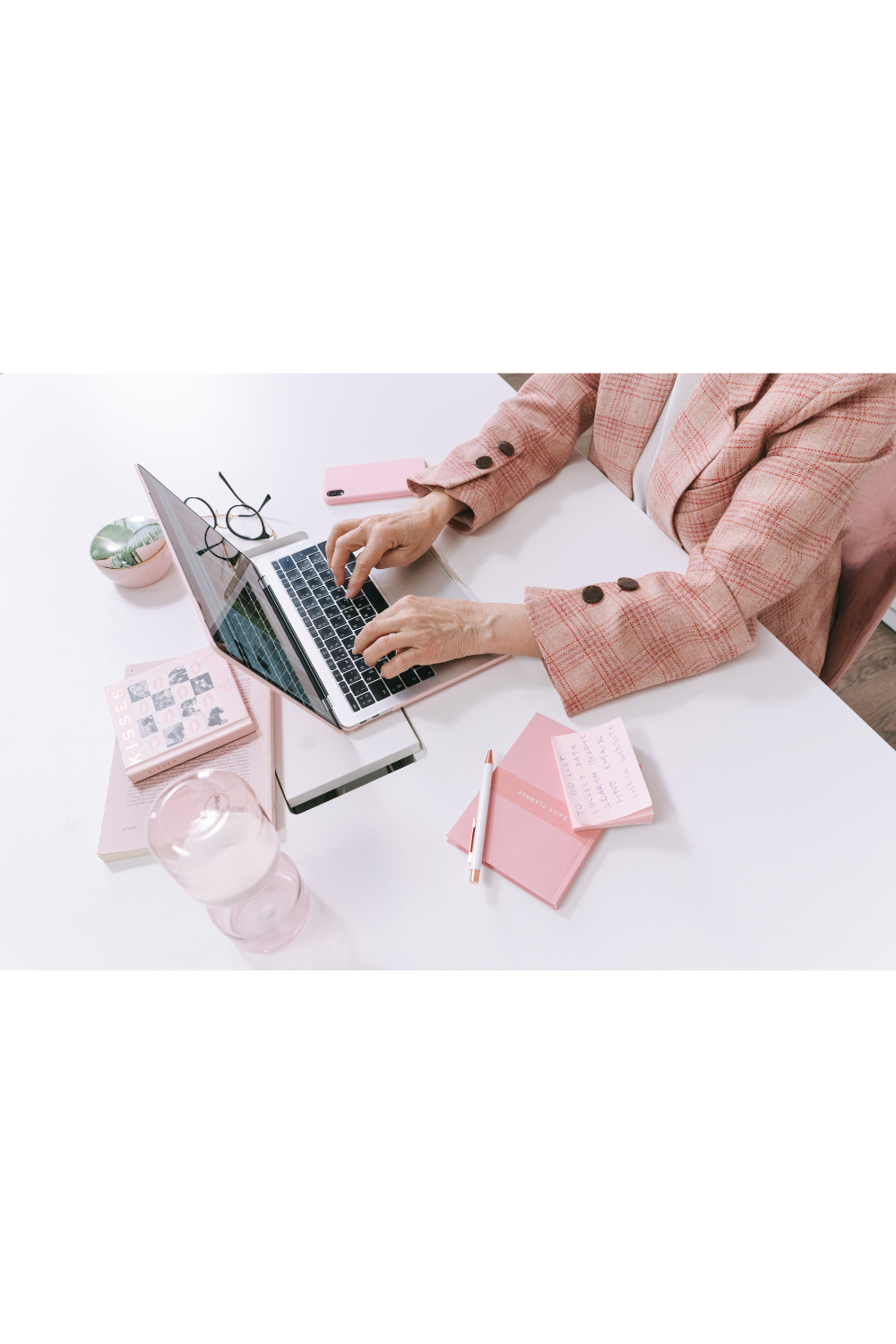 A person using a laptop at a white desk with pink and green accessories, glasses, a pink phone, notebooks, a pen, and a glass of water.
