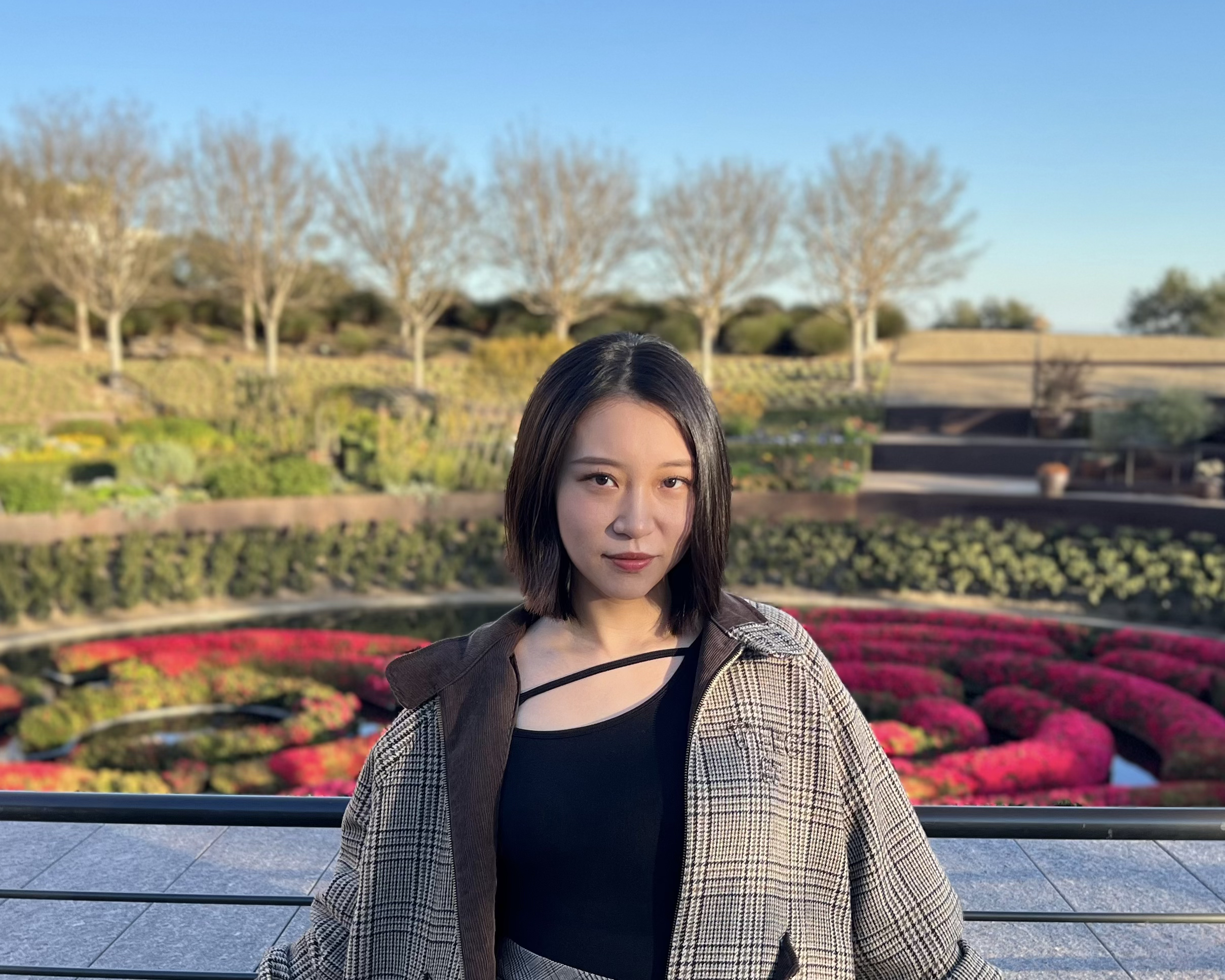 Young woman with shoulder-length dark hair standing outdoors near a railing, with trees and flower beds in the background on a sunny day.