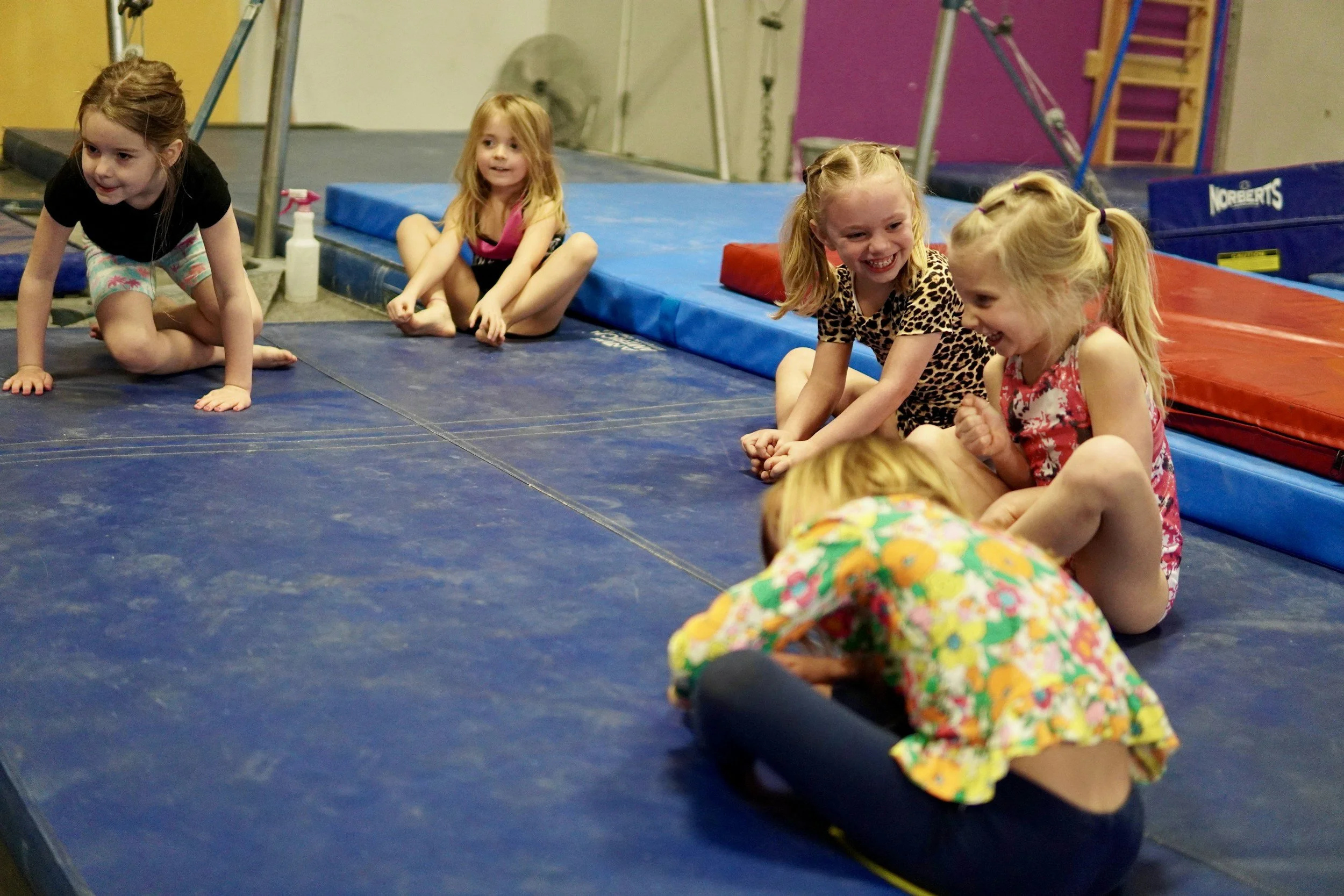Four young girls playing and laughing on gymnastics mats in a gym. One girl is crawling with her hands and knees, while the others are sitting and smiling.