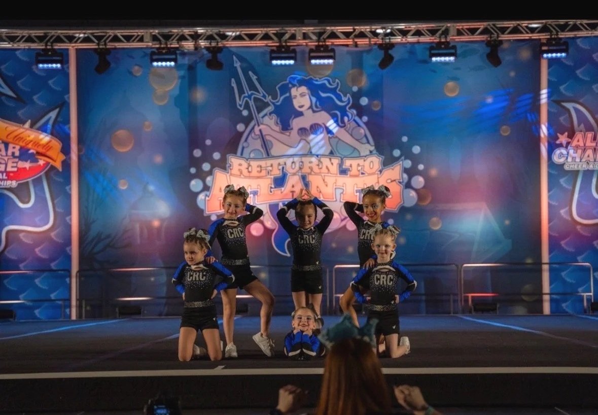 Cheerleader team in blue and black uniforms performing a routine on stage at a cheerleading competition, with a colorful backdrop featuring a mermaid illustration and the words 'Born to Perform'.