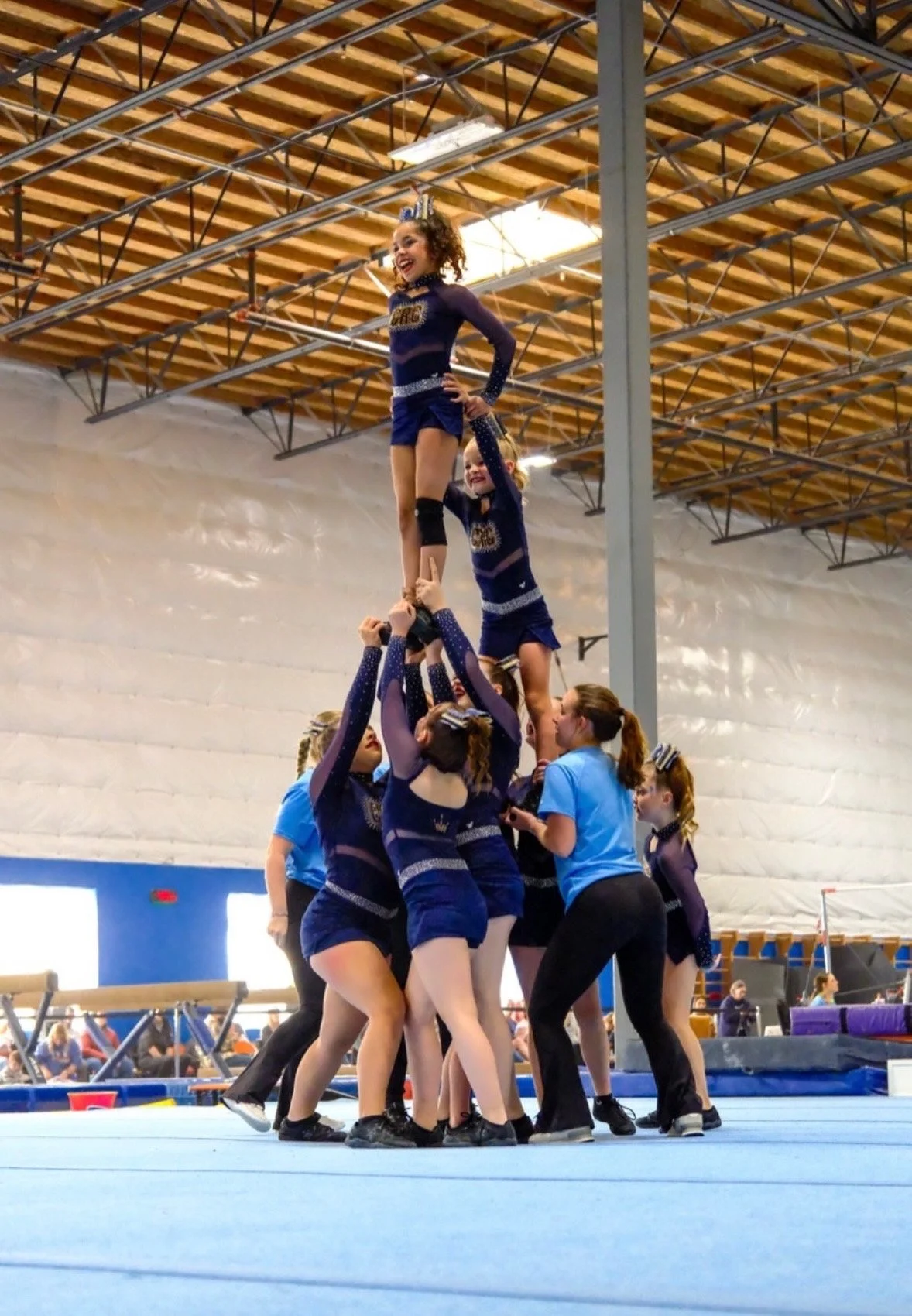 Cheerleaders forming a pyramid in a gymnasium, with one girl at the top smiling, while others support her from below. The scene includes coaches and team members watching and guiding.