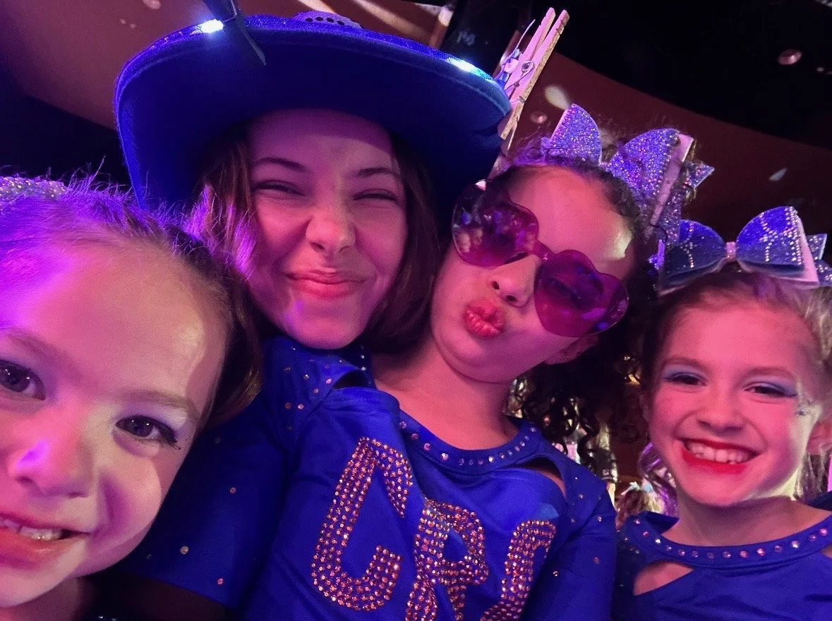 Four young girls, dressed in blue with glitter and accessories, smiling and making playful faces at a party.