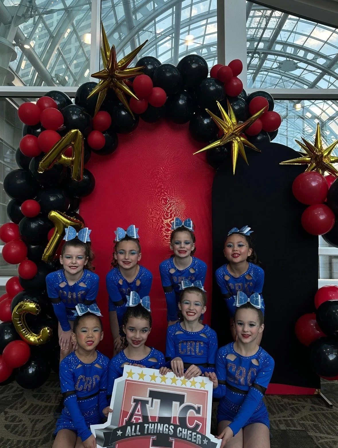 Group of cheerleaders in blue costumes with bows, posing in front of a red and black balloon arch decorated with gold stars, at a cheerleading event.