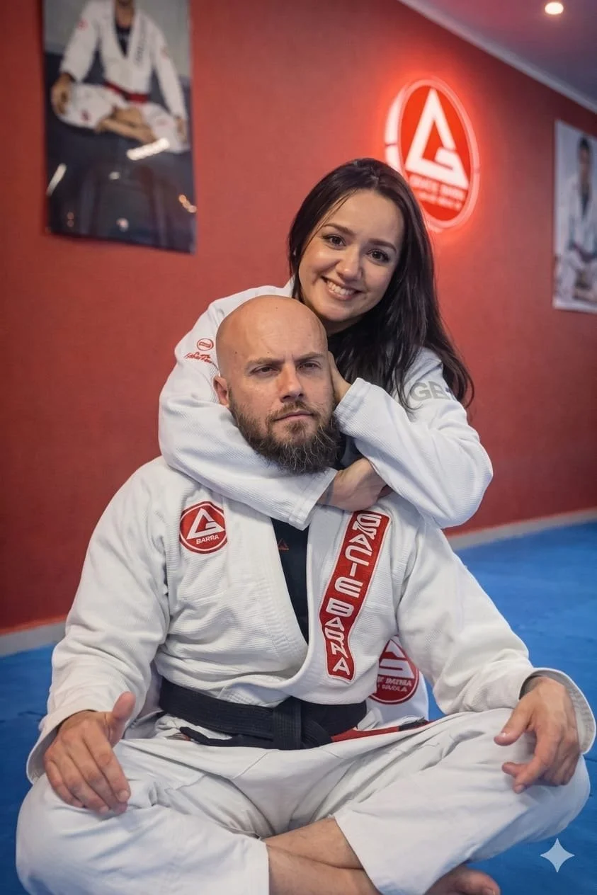 A woman in a martial arts gi smiling and hugging a man in a martial arts gi, who is seated cross-legged on a blue mat in a martial arts studio with red walls and martial arts posters.