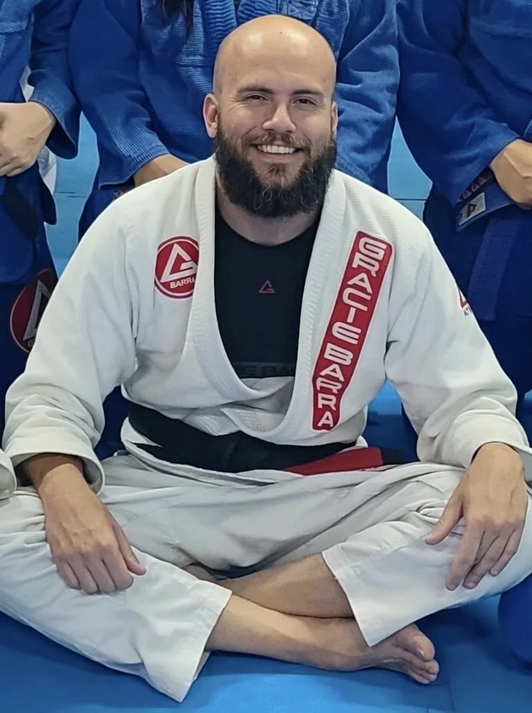 Man with beard and bald head smiling, sitting cross-legged on a blue mat, wearing a white Brazilian Jiu-Jitsu gi with red patches, surrounded by others in gi uniforms.