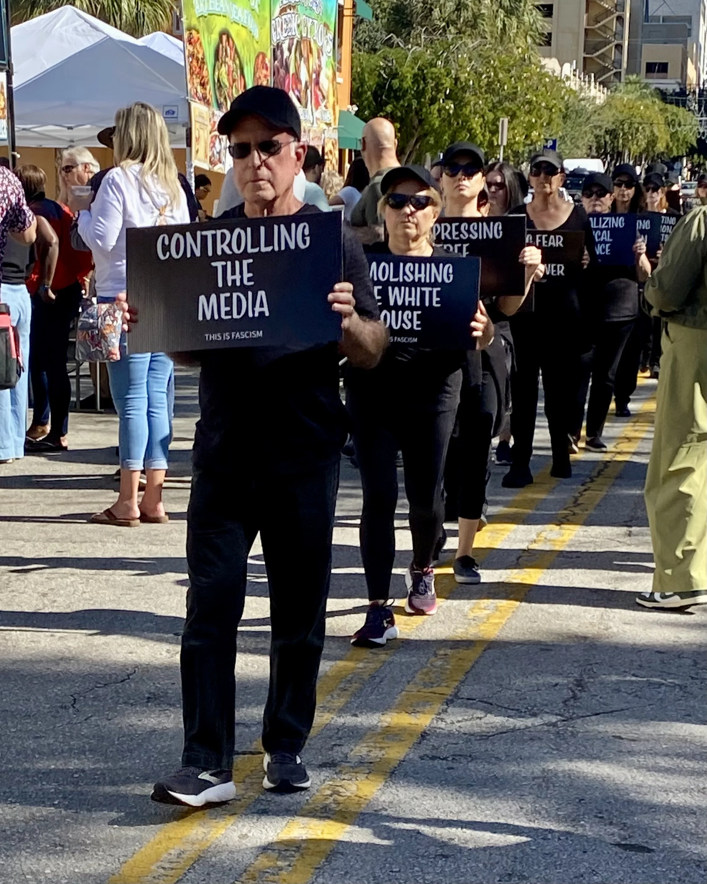 A group of people participating in a protest or march, holding signs with messages like 'Controlling the Media.' The marchers are walking on a city street lined with trees, tents, and colorful signs, with buildings in the background.